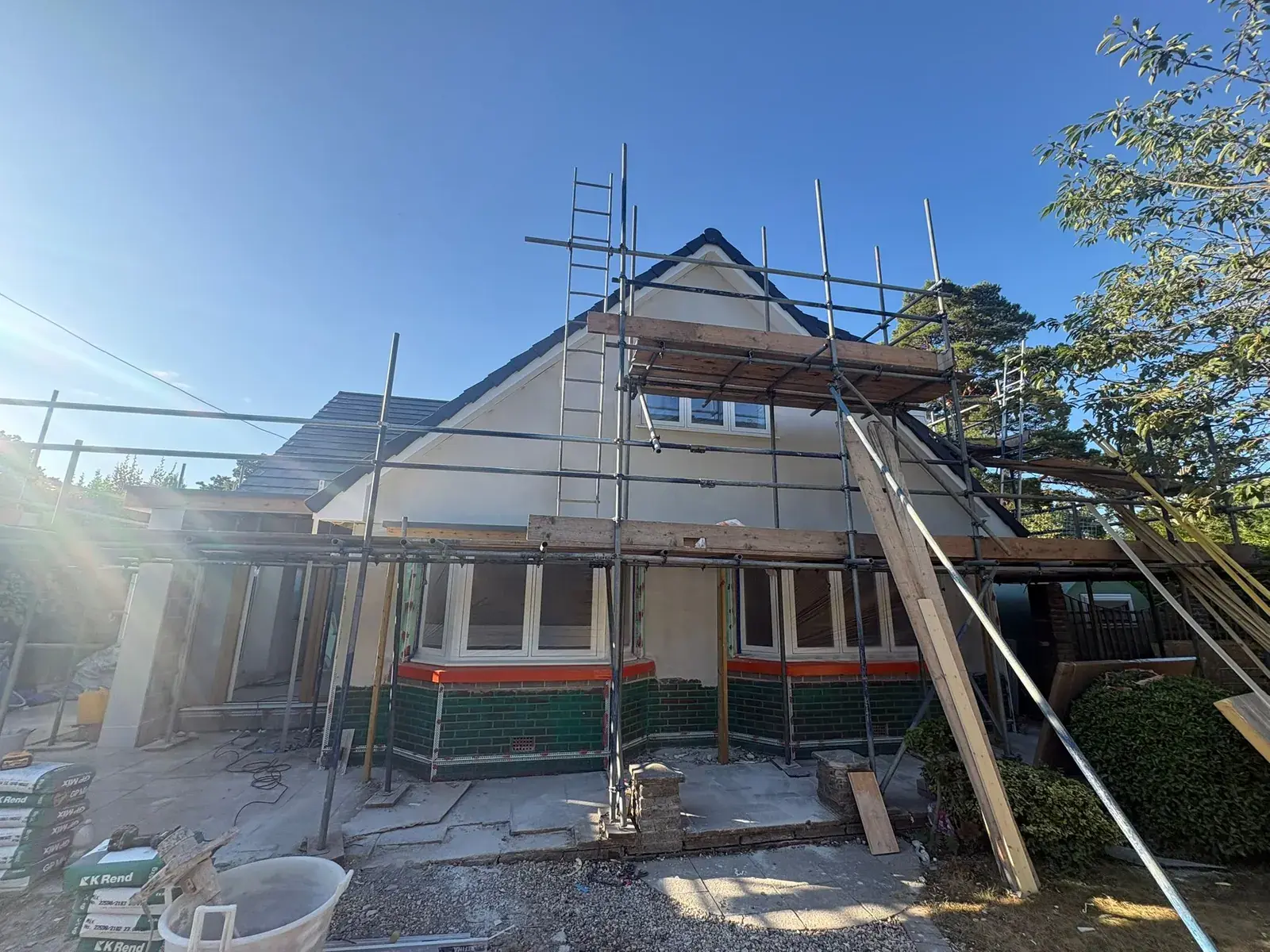 Chalet bungalow on scaffold with green tiled bay base, K Rend bags and mixing tub on the drive