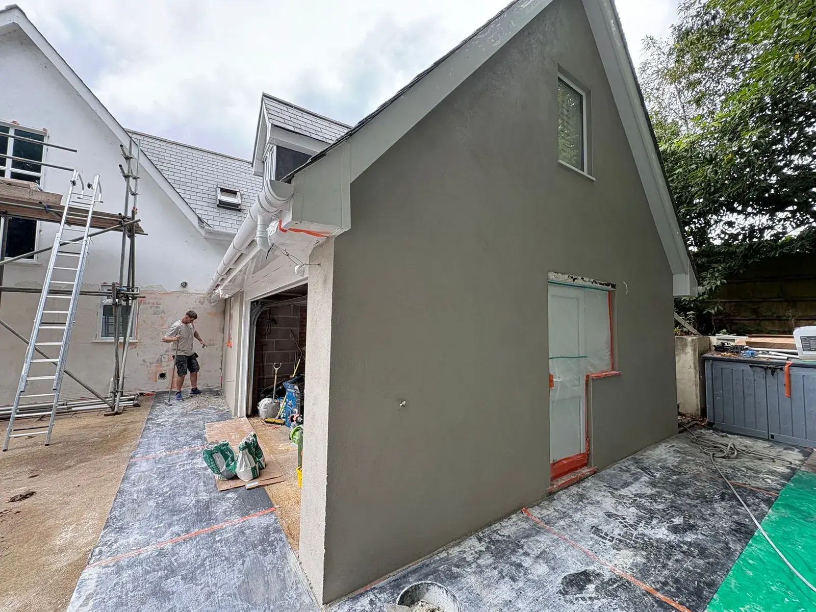 Gable end of an extension in fresh grey base coat, plasterer working on the main house wall behind