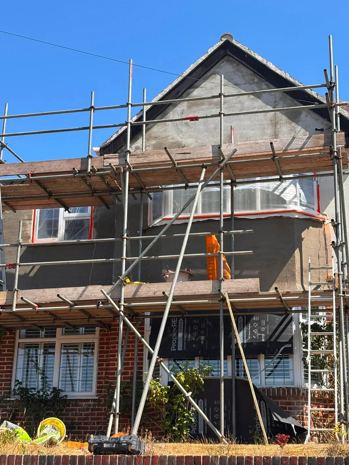1930s house on scaffold with mesh and base coat over the upper floor, windows taped, brick ground floor sheeted in black protection