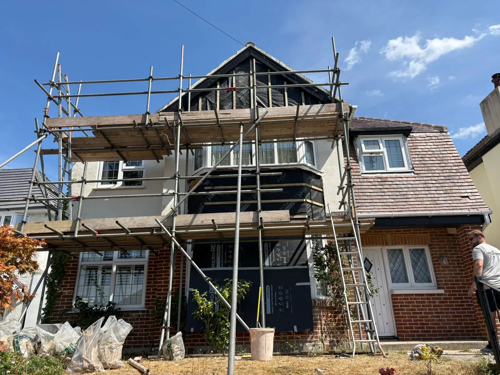 Detached house on scaffold with windows and brickwork sheeted in black protection, bags of rubble on the lawn