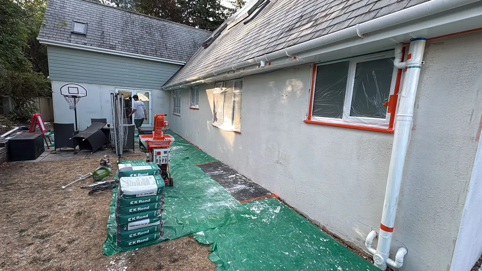 Render spray pump and a stack of K Rend bags on green sheeting beside a house with the base coat going on